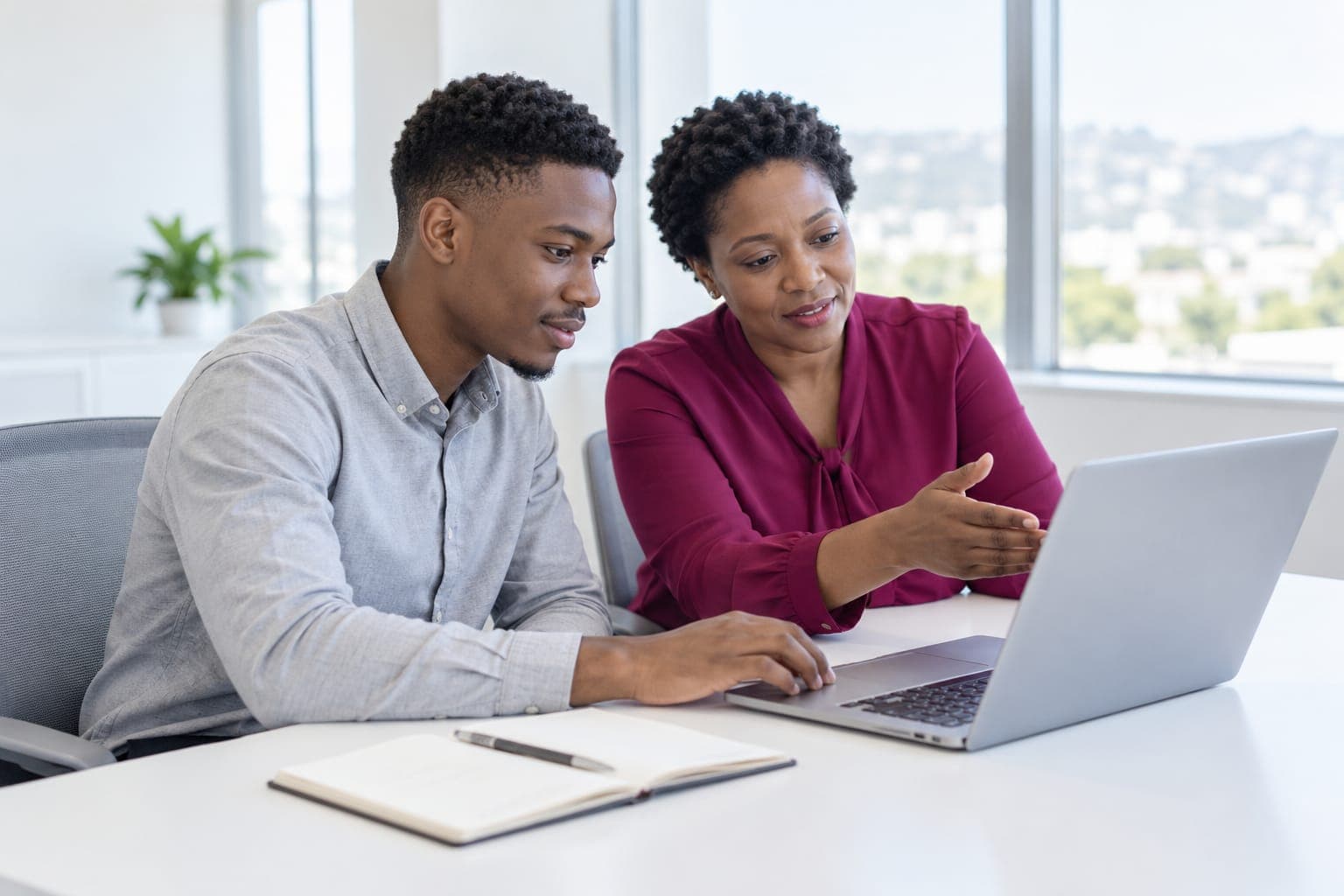 Supervisor in a burgundy blouse guiding a young intern through work on a laptop