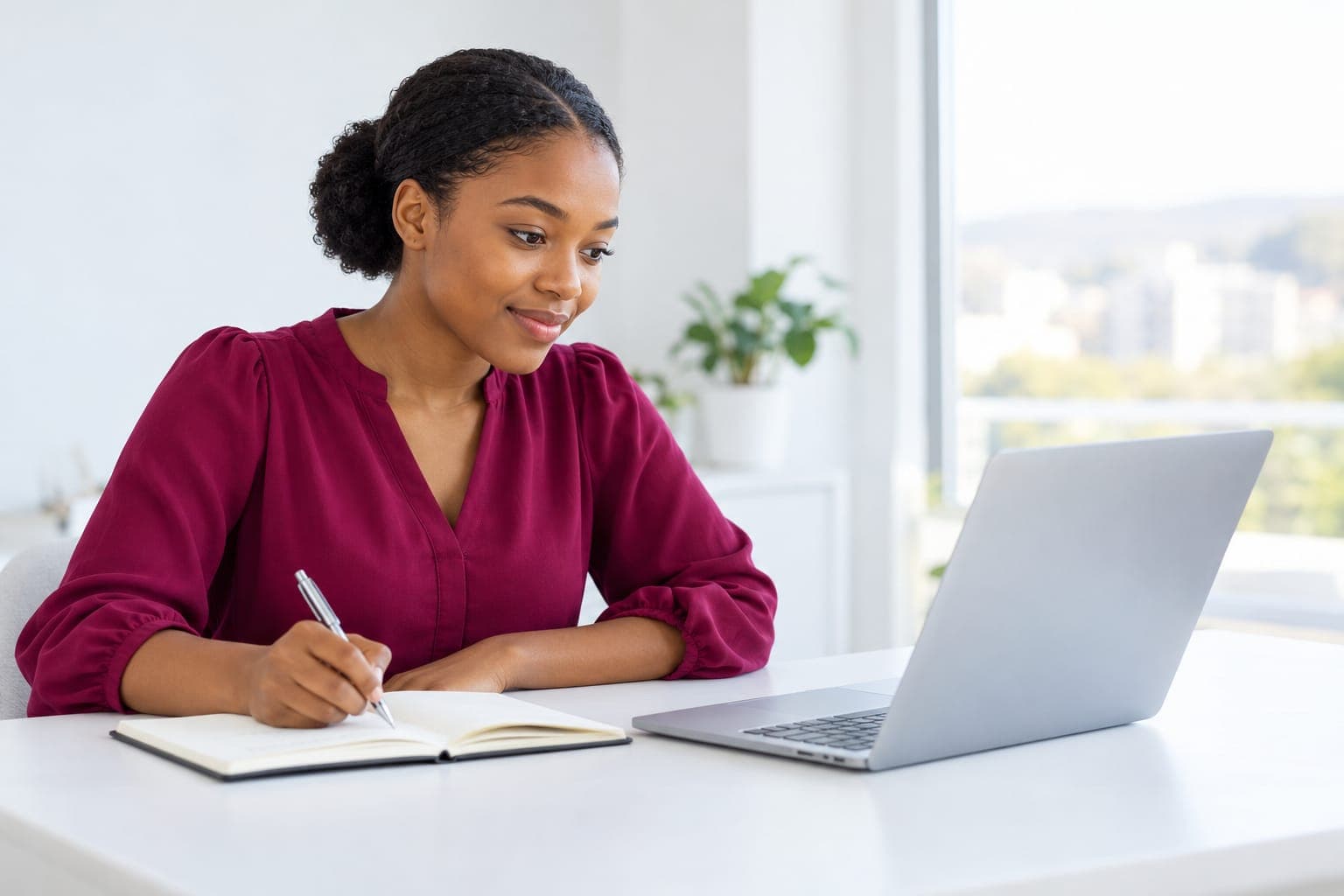 Young woman in a burgundy blouse smiling as she takes notes beside her laptop